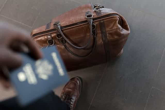 Man holding a passport, standing next to a brown travel bag